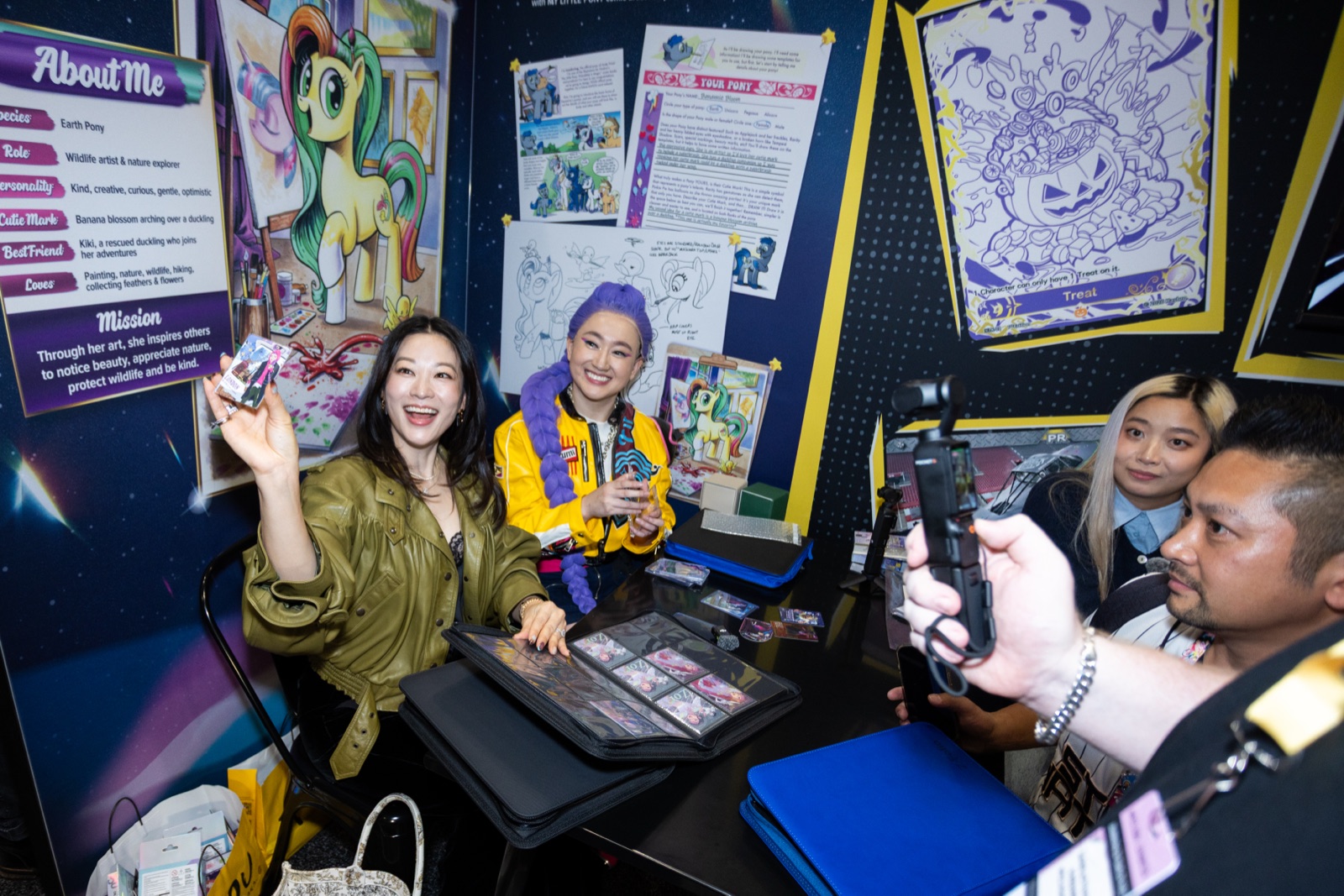 Arden Cho holds up a KAYOU card at a booth table covered in binders, with fans across from her